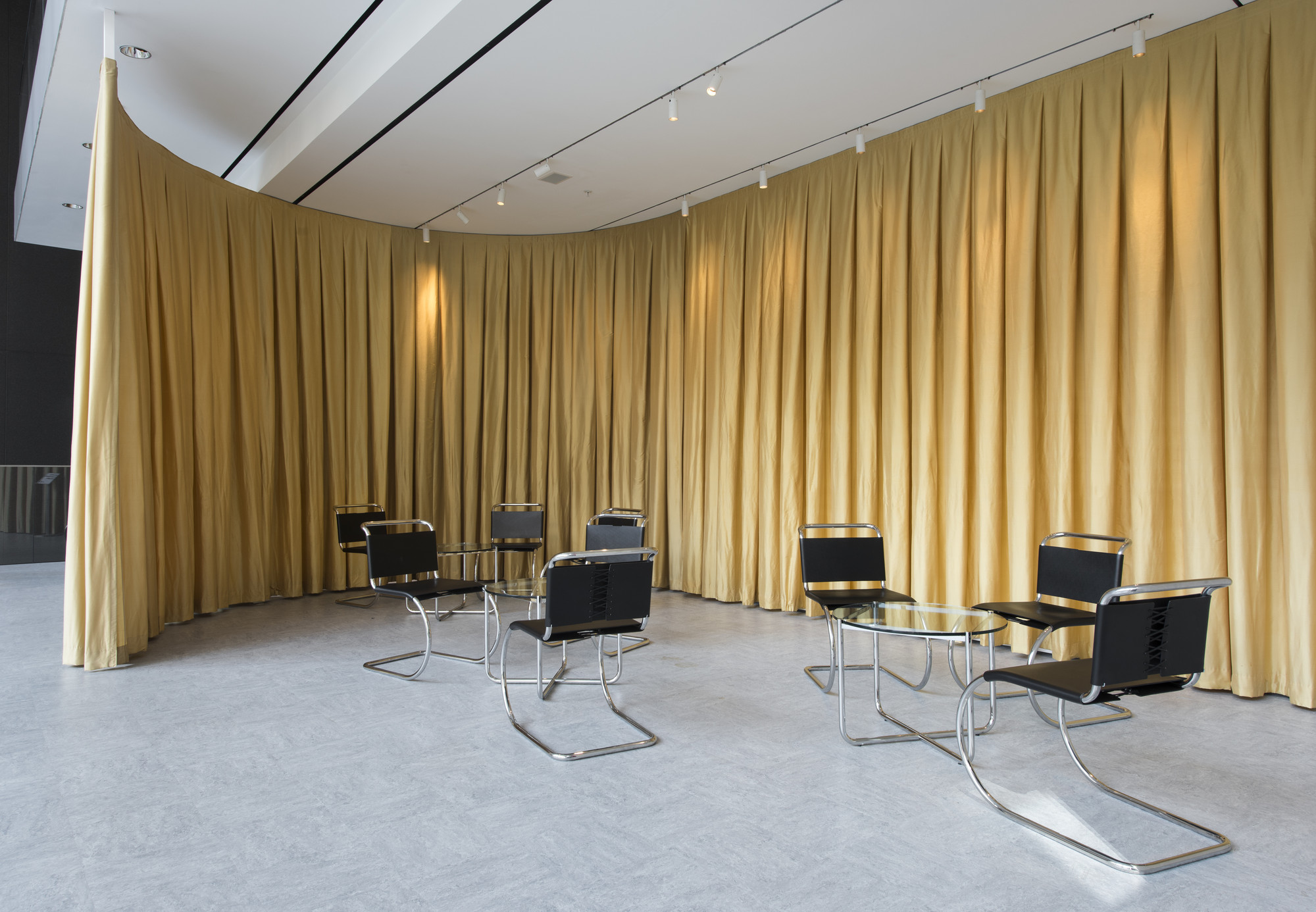 A curved golden curtain behind black cantilevered tubular steel chairs and small round glass tables in a reconstruction of the Velvet and Silk Cafe